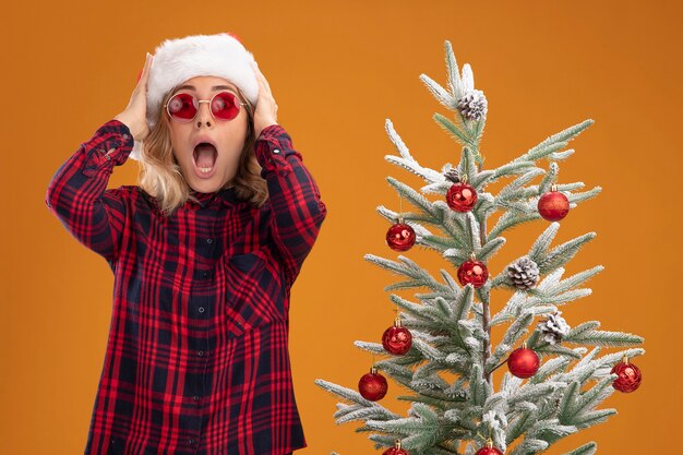 Erschrockenes junges schönes Mädchen, das in der Nähe des Weihnachtsbaums mit Weihnachtsmütze mit Brille steht, packte den Kopf isoliert auf orangem Hintergrund