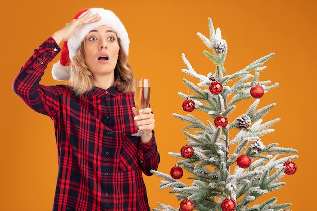Kostenloses Foto erschrockenes junges schönes mädchen, das in der nähe des weihnachtsbaums steht und einen weihnachtshut trägt, der ein glas champagner hält und mit der hand isoliert auf orangefarbenem hintergrund schaut