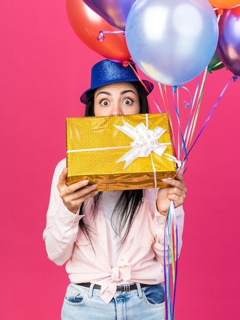 Erschrockenes junges schönes Mädchen mit Partyhut mit Luftballons und bedecktem Gesicht mit Geschenkbox isoliert auf rosa Wand