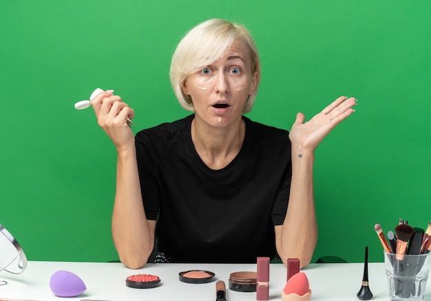 Erschrockenes junges schönes Mädchen sitzt am Tisch mit Make-up-Tools, die Ton-up-Creme auftragen, die Hände einzeln auf grünem Hintergrund ausbreitet