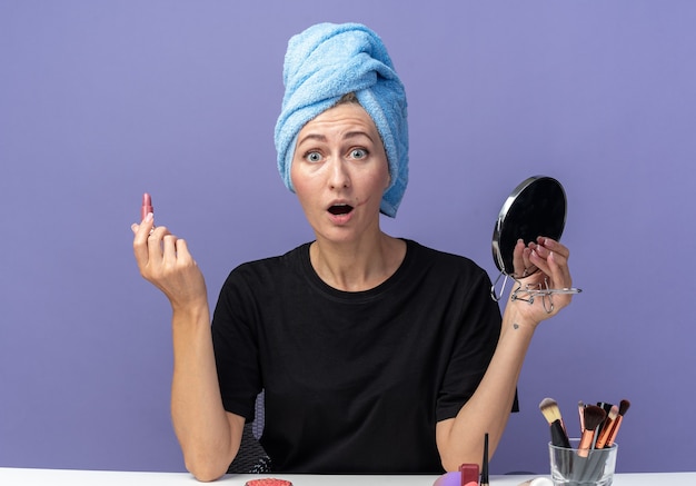 Erschrockenes junges schönes Mädchen sitzt am Tisch mit Make-up-Tools und wischt sich die Haare im Handtuch ab und hält Lippenstift mit Spiegel isoliert auf blauem Hintergrund