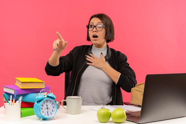 Erschrockenes junges Studentenmädchen, das eine Brille trägt, die am Schreibtisch sitzt und Hausaufgaben macht und zur Seite mit der Hand auf der Brust zeigt, die auf rosa Wand lokalisiert wird