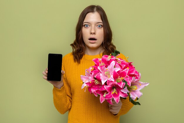 Erschrockenes schönes junges Mädchen am Tag der glücklichen Frau, das Blumenstrauß mit Telefon lokalisiert auf olivgrüner Wand hält