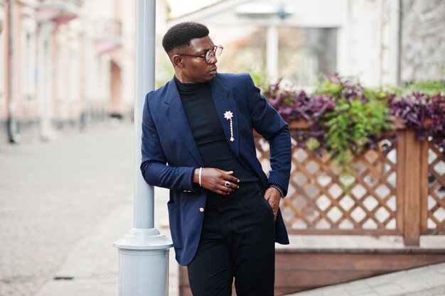 Erstaunlich aussehender afrikanisch-amerikanischer Mann trägt einen blauen Blazer mit Brosche, schwarzem Rollkragenpullover und Brille, die auf der Straße gestellt wurden Modischer schwarzer Typ