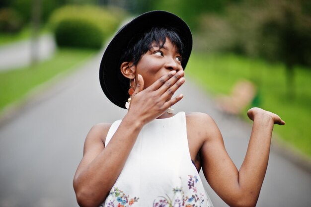 Erstaunliche afroamerikanische Modelfrau in grüner Hose und schwarzem Hut, die mit unterschiedlichen Emotionen im Park posiert, zeigt ein überraschendes Gesicht