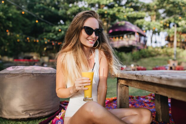 Erstaunliche blondhaarige Frau in der Sonnenbrille, die Orangencocktail im Park trinkt.