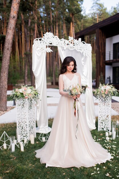 Erstaunliche Braut in einem beige Kleid steht vor einem Hochzeitsaltar draußen
