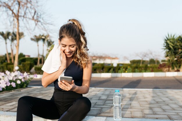 Erstaunliche freudige hübsche junge Frau in Sportbekleidung, mit langen lockigen Haaren, die am Morgen draußen im Park am Telefon plaudern. Training, fröhliche Stimmung, Glück, Sonnenaufgang, Freude, gesunder Lebensstil