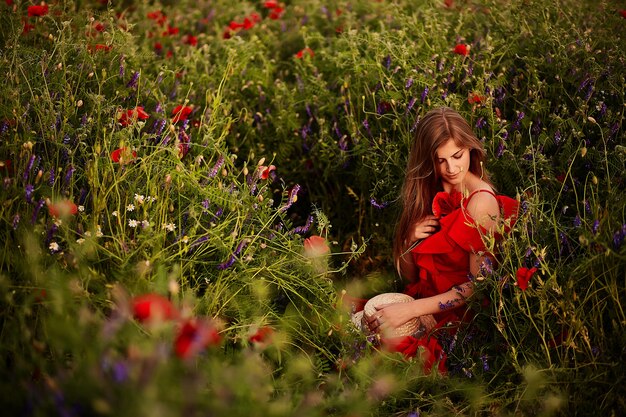 Erstaunliche junge Frau im roten Kleid sitzt auf dem grünen Feld mit roten Mohnblumen