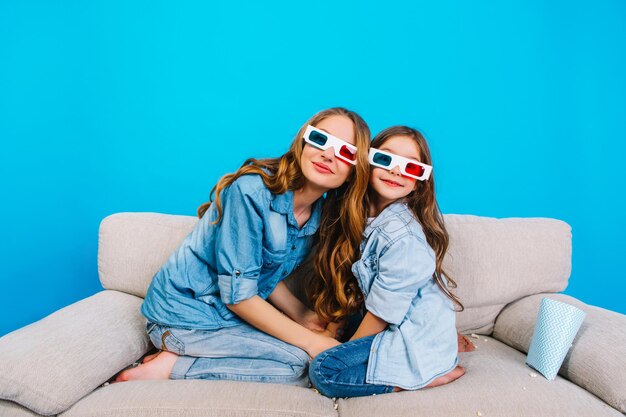 Erstaunliche Mutter und Tochter, die sich in 3D-Brille umarmen und lächeln, um die Kamera auf der Couch einzeln auf blauem Hintergrund zu fotografieren. Wahre glückliche Emotionen ausdrücken, Familienzeit der Mutter mit süßem Kind, gemeinsame Unterhaltung