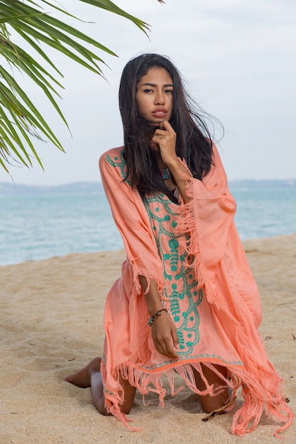 Erstaunliche sexy tan asiatische Frau, die auf tropischem Strand des Paradieses unter dem pam Baum aufwirft, auf weißem Sand sitzt, sich entspannt und Feiertage genießt. Boho Kleid mit Stickerei. Bali.