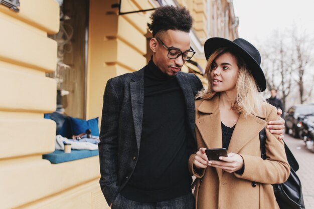 Erstaunliches blondes Mädchen, das mit Interesse auf ihren afrikanischen Freund schaut. Porträt im Freien von jungen Leuten im trendigen Outfit, die Zeit zusammen verbringen.