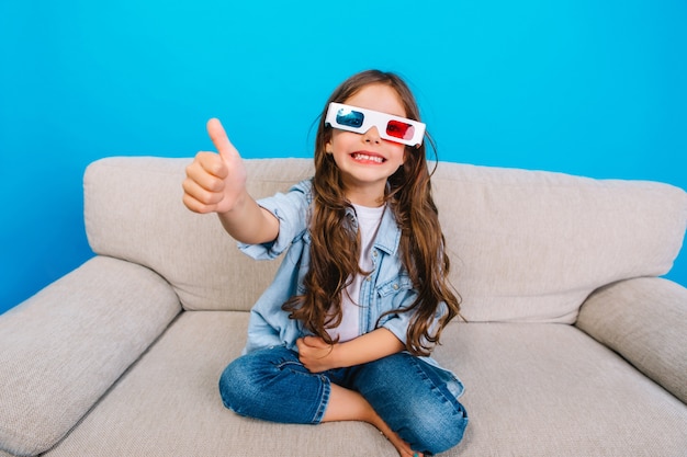 Erstaunliches glückliches kleines Mädchen in der 3D-Brille mit dem langen brünetten Haar, das zur Kamera auf der Couch lächelt, lokalisiert auf blauem Hintergrund. Zeigt wahre positive Emotionen, glückliche Kindheit des modischen Kindes