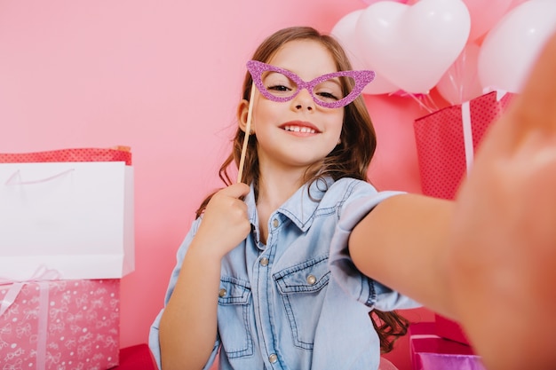 Erstaunliches kleines Mädchen des Selbstporträts mit lila Maske auf Gesicht, das zur Kamera auf rosa Hintergrund lächelt. Feiern Sie alles Gute zum Geburtstag, bunte Luftballons mit großen Geschenkboxen, die Positivität ausdrücken