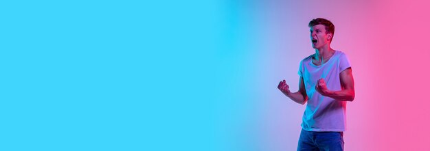 Erstaunt. Porträt des jungen kaukasischen Mannes auf blau-rosa Studiohintergrund des Gradienten im Neonlicht. Jugendkonzept, menschliche Emotionen, Gesichtsausdruck, Verkauf, Werbung. Schönes Modell in lässig. Flyer