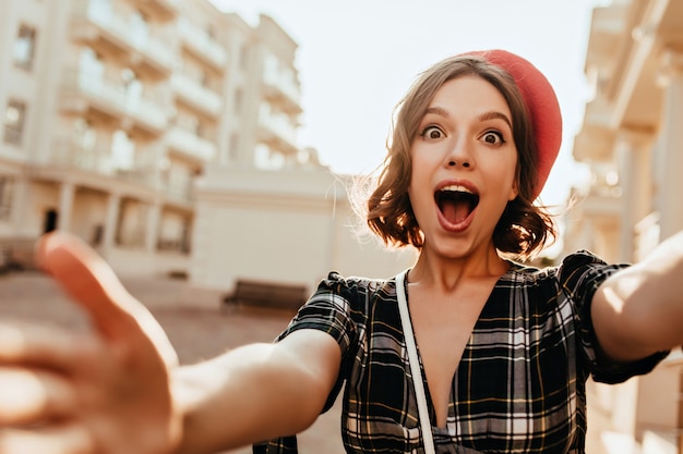 Erstaunte französische Dame mit braunen Augen, die Selfie auf der Straße machen. Lustiges lockiges Mädchen in der roten Baskenmütze, die Foto von sich selbst beim Gehen durch Stadt macht.