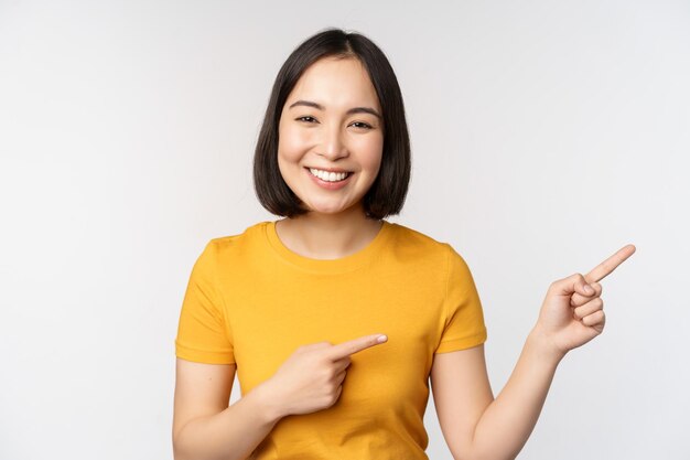 Erstaunte junge Asiatin, die Werbung beiseite zeigt und mit dem Finger direkt auf das Markenlogo des Werbetextes zeigt, das glücklich vor weißem Hintergrund steht