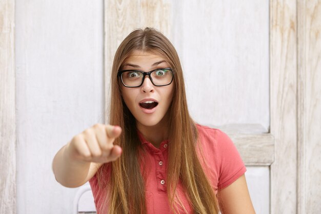 Erstaunte junge Frau mit rechteckiger Brille und Poloshirt, die ihren Zeigefinger nach vorne zeigt