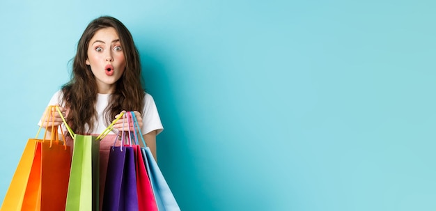 Erstaunte junge Frau Shopaholic hält bunte Einkaufstaschen und schaut amüsiert auf den nächsten Laden, der Thi kauft