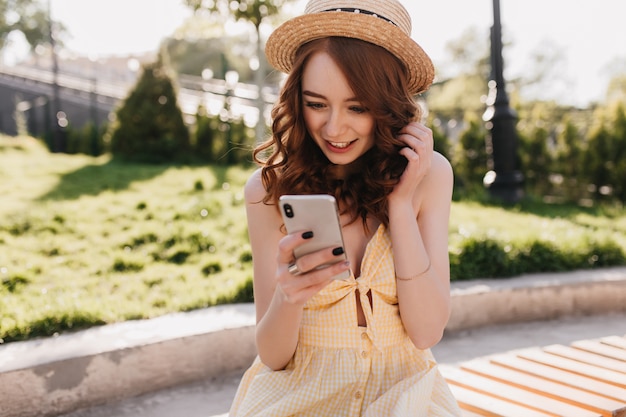 Erstaunte junge Ingwerfrau las telefonische Nachricht im Park. Außenporträt des reizenden eleganten Mädchens im gelben Kleid, das auf Bank mit Smartphone sitzt.