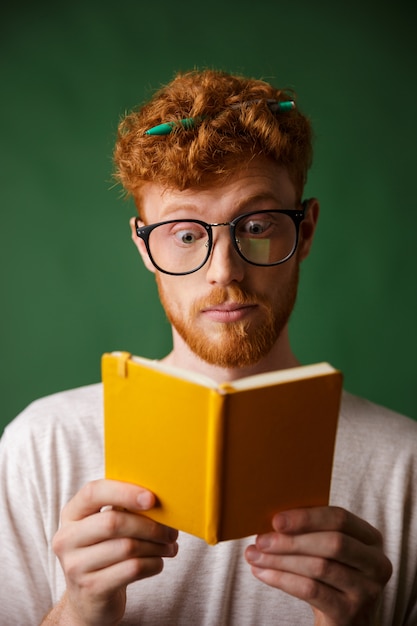 Erstaunter junger roter bärtiger Student in der Brille, der das Notizbuch mit Stift in seinen Haaren liest