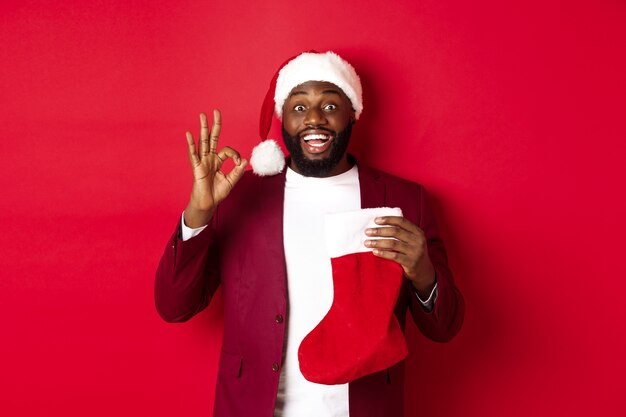 Erstaunter schwarzer Mann, der Weihnachtsgeschenke in der Weihnachtssocke hält und ein OK-Zeichen zeigt, wie etwas, das über rotem Hintergrund steht
