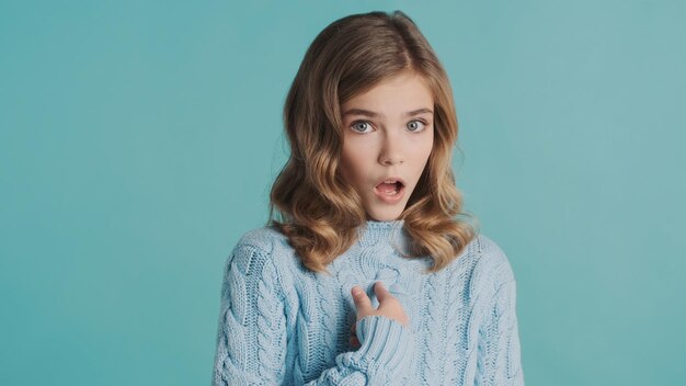 Erstauntes blondes Teenager-Mädchen im Pullover, das mit den Fingern auf sich selbst zeigt und schockiert vor der Kamera über blauem Hintergrund aussieht