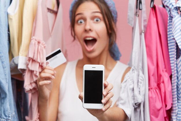 Erstauntes weibliches Model, das mit abgehörten Augen und weit geöffnetem Mund schaut, während Kreditkarte in einer Hand und Smartphone mit leerem Bildschirm in der anderen Hand in ihrer Garderobe stehen