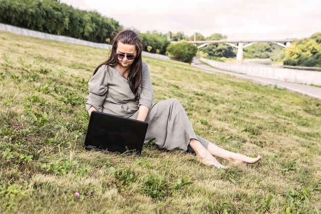 Erwachsene frau sitzt auf dem gras im park und arbeitet an einem laptop
