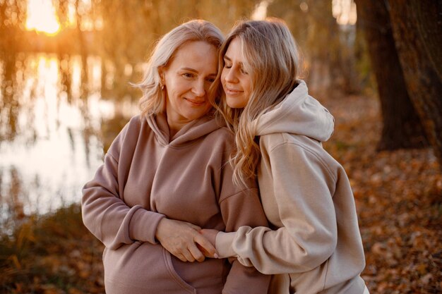 Erwachsene Tochter umarmt ihre schwangere Mutter an einem sonnigen Herbsttag in der Natur Blonde Frauen stehen im Park in der Nähe des Sees Frauen tragen beige Kleidung