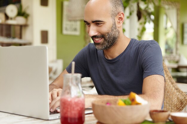Erwachsener bärtiger Mann, der im Café frühstückt, am Tisch vor dem allgemeinen Laptop sitzt und mit dem Lächeln schaut, während er Freunde über soziale Netzwerke mitteilt und kostenloses Wi-Fi genießt.