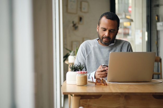 Erwachsener Geschäftsmann im Pullover nutzt Smartphone aufmerksam, während er im Stadtcafé am Laptop arbeitet