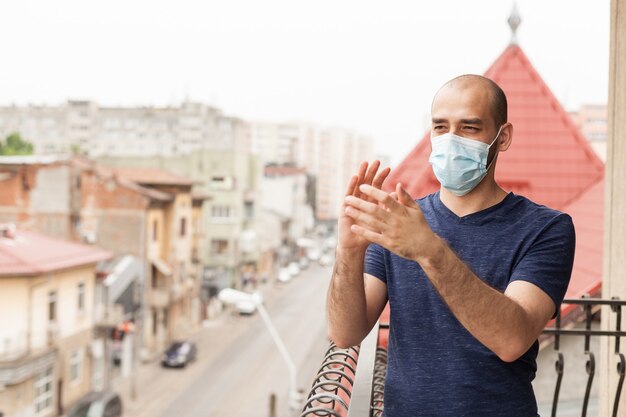 Erwachsener Mann mit Schutzmaske, der auf Balkon klatscht und Unterstützung für medizinisches Personal im Kampf mit Coronavirus zeigt.