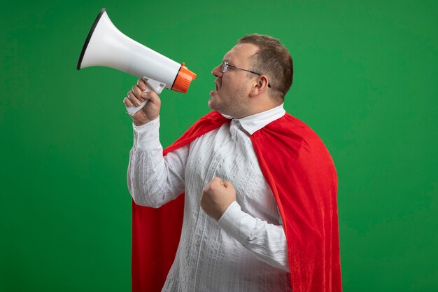 Erwachsener slawischer Superheldenmann im roten Umhang, der die Brille trägt, die in der Profilansicht steht, die durch die geballte Faust des Lautsprechers spricht, die auf grüner Wand mit Kopienraum isoliert wird
