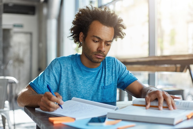 Kostenloses Foto erwachsener student, der drinnen in einem geräumigen raum sitzt, der feierlichen ausdruck sieht, der in bücher und notizbuch schaut, etwas schreibt, während er sich für klassen vorbereitet. studien- und lernkonzept