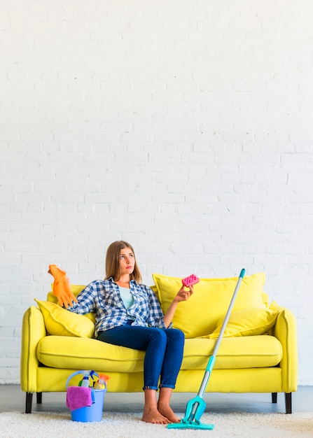 Erwogene junge Frau, die auf dem gelben Sofa hält Schwamm- und Gummihandschuhe sitzt