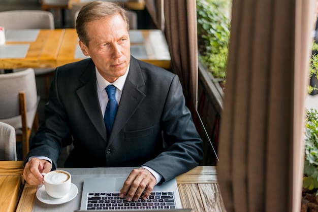 Erwogener Geschäftsmann, der heraus durch Glasfenster mit Tasse Kaffee und Laptop auf Schreibtisch schaut