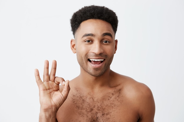 Es in Ordnung. Mach dir keine Sorgen. Nahaufnahme des fröhlichen jungen attraktiven dunkelhäutigen Mannes mit Afro-Frisur mit nacktem Oberkörper, der OK-Zeichen mit Hand zeigt, zwinkert und in der Kamera mit flirtendem Ausdruck schaut.