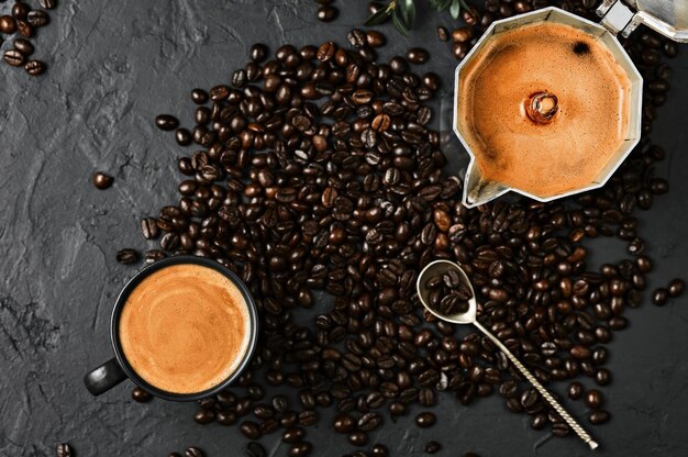 Espressokaffee mit duftendem Schaum in einer Tasse und in einer Kanne und Kaffeebohnen auf einem schwarzen Tisch, flach gelegt. Frühstück mit italienischem Café, Draufsicht der Kaffeekanne auf dem Tisch