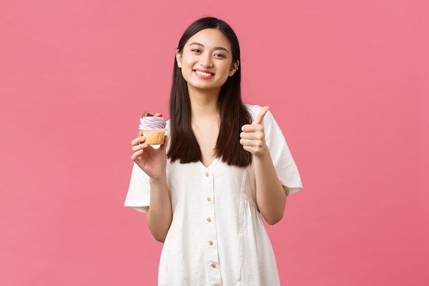 Essen, Café und Restaurants, Sommer-Lifestyle-Konzept. Fröhliche asiatische Frau, die mit fantastischem Dessertgeschmack zufrieden ist, Daumen hoch als Empfehlung Cupcake aus der Bäckerei, stehend rosa Hintergrund.