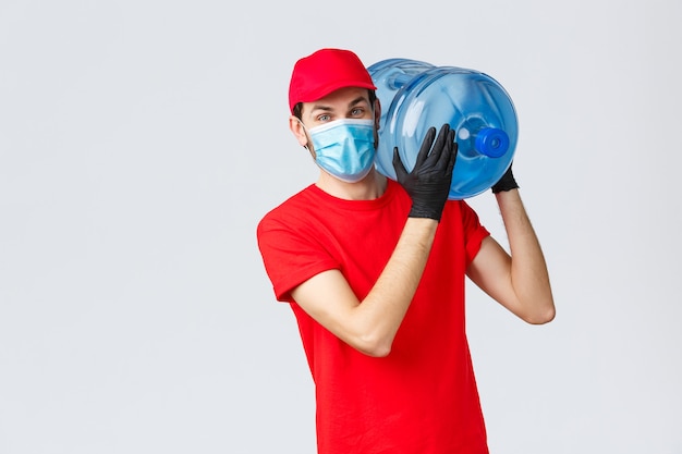 Essen zum Mitnehmen, Lieferung von Lebensmitteln und Lebensmitteln, Konzept für kontaktlose Bestellungen von Covid-19. Freundlicher Kurier in roter Uniform, Mütze, Gesichtsmaske und Handschuhen, der eine Wasserflaschenbestellung auf der Schulter hält