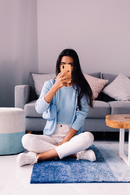 Europäische fit brünette Modebloggerin sitzt auf dem Boden im Wohnzimmer nahe Sofa mit Telefon