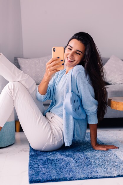 Europäische fit brünette Modebloggerin sitzt auf dem Boden im Wohnzimmer nahe Sofa mit Telefon