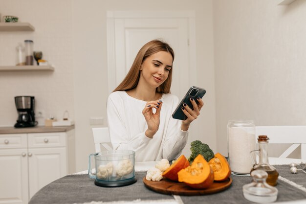 Europäische frau mit hellbraunem haar lacht und sucht rezept im smartphone. junge dame, die sich zum kochen mit kürbis und brokkoli vorbereitet,