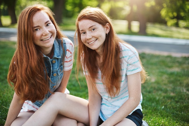 Europäische Schwestern mit roten Haaren und Sommersprossen sitzen auf grünem Gras und lächeln breit, hängen mit Freunden beim Picknick ab und drücken Freude und Vergnügen aus. Emotionen und Familienkonzept