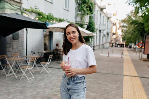 Europäische stilvolle Frau mit kurzer dunkler Frisur in weißem T-Shirt und Jeans, die mit wunderbarem Lächeln vor der Kamera posiert und Smoothie auf der Sommerstraße von Sunne hält