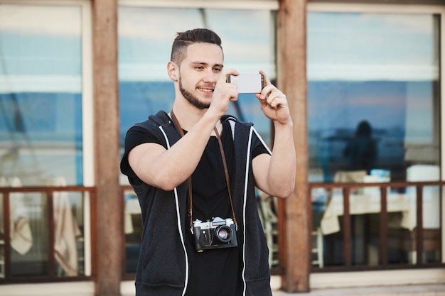 Europäischer Fotograf in trendiger Kleidung, die Foto am Telefon beim Tragen der Kamera am Hals macht