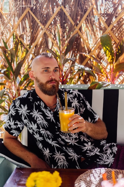 Europäischer hübscher bärtiger Mann trinkt Mango-Smoothie im Sommercafé, tropische Wand dahinter.