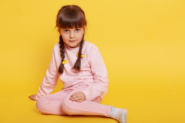 Europäisches entzückendes Mädchen, das auf Boden sitzt und blassrosa Hosen und Pullover trägt, lokalisiert über gelbem Hintergrund, schaut auf Kamera, dunkelhaariges weibliches Kind mit Zöpfen.