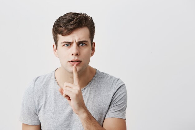 Europäisches männliches Model in grauem T-Shirt mit Zeigefinger auf den Lippen, Stirnrunzeln im Gesicht, Bitte um Zunge und vertrauliche Informationen. Streng geheim